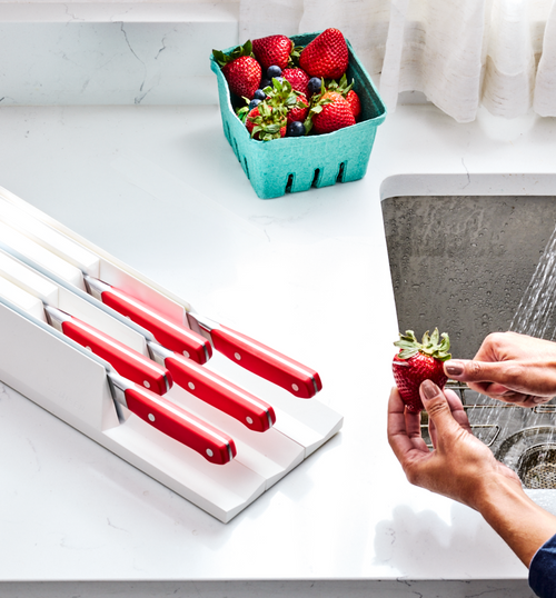 Red-handled kitchen knives in white knife block with fresh strawberries being rinsed at sink