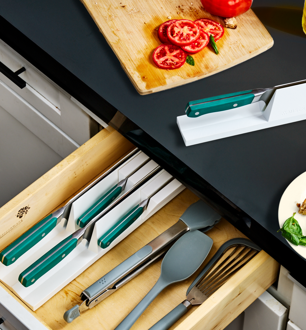 Kitchen drawer knife organizer with teal-handled knives, spatulas, and slotted spoon beside cutting board with sliced tomatoes